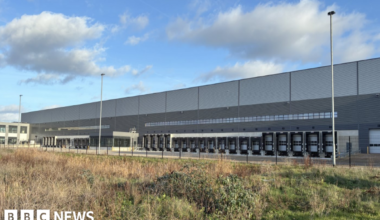 A shot of the new distribution centre - it is a large grey rectangular building with multiple lorry loading bays on the side and office buildings to the left