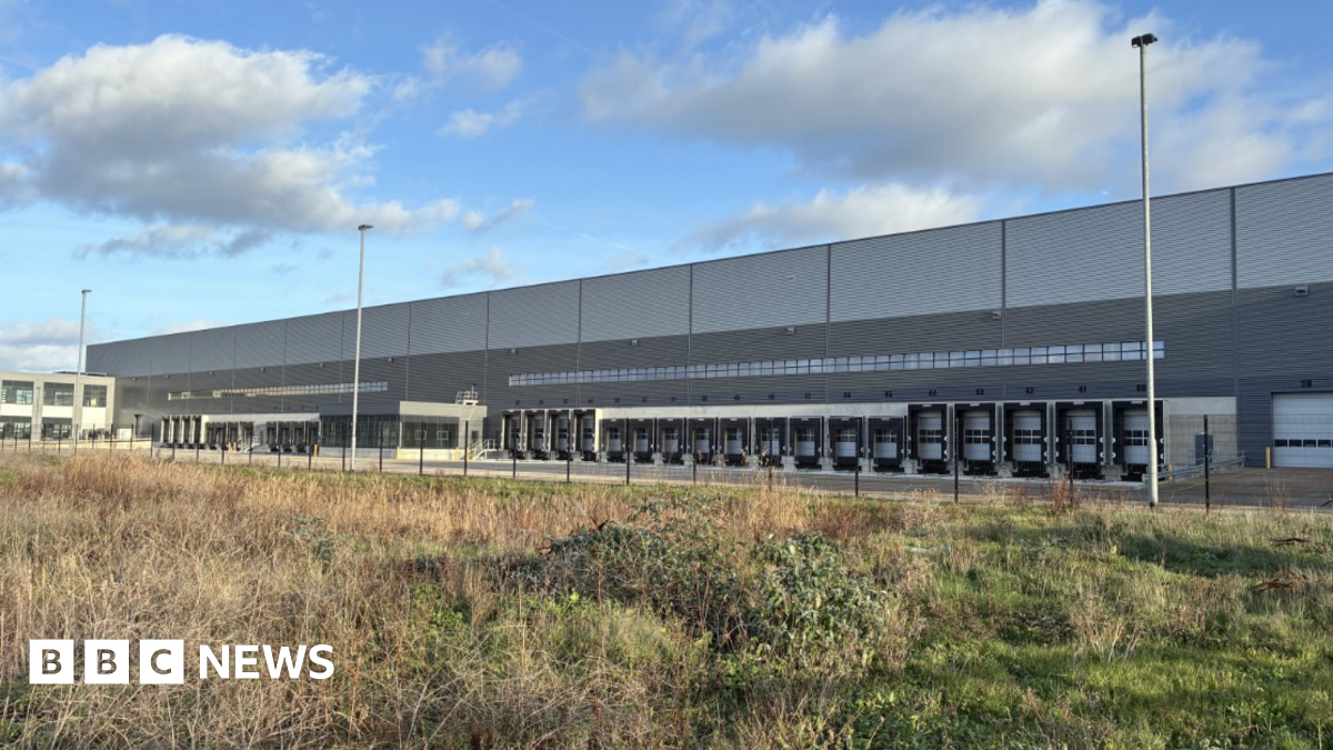 A shot of the new distribution centre - it is a large grey rectangular building with multiple lorry loading bays on the side and office buildings to the left