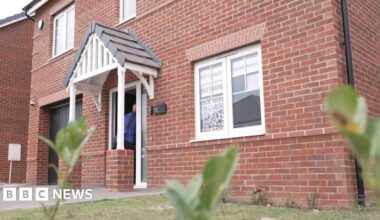 A newbuild red-brick detached house. It has white window frames. In the window closest to the camera are signs reading: Top quality Avant Homes not mine See why...
