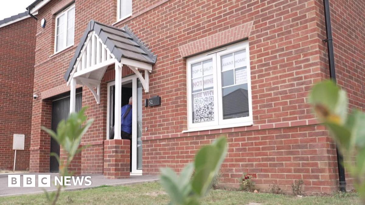 A newbuild red-brick detached house. It has white window frames. In the window closest to the camera are signs reading: Top quality Avant Homes not mine See why...