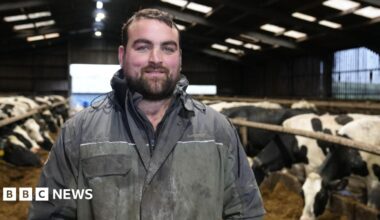 Gethin Hughes, dairy farmer stands in his cattle shed wearing blue overalls