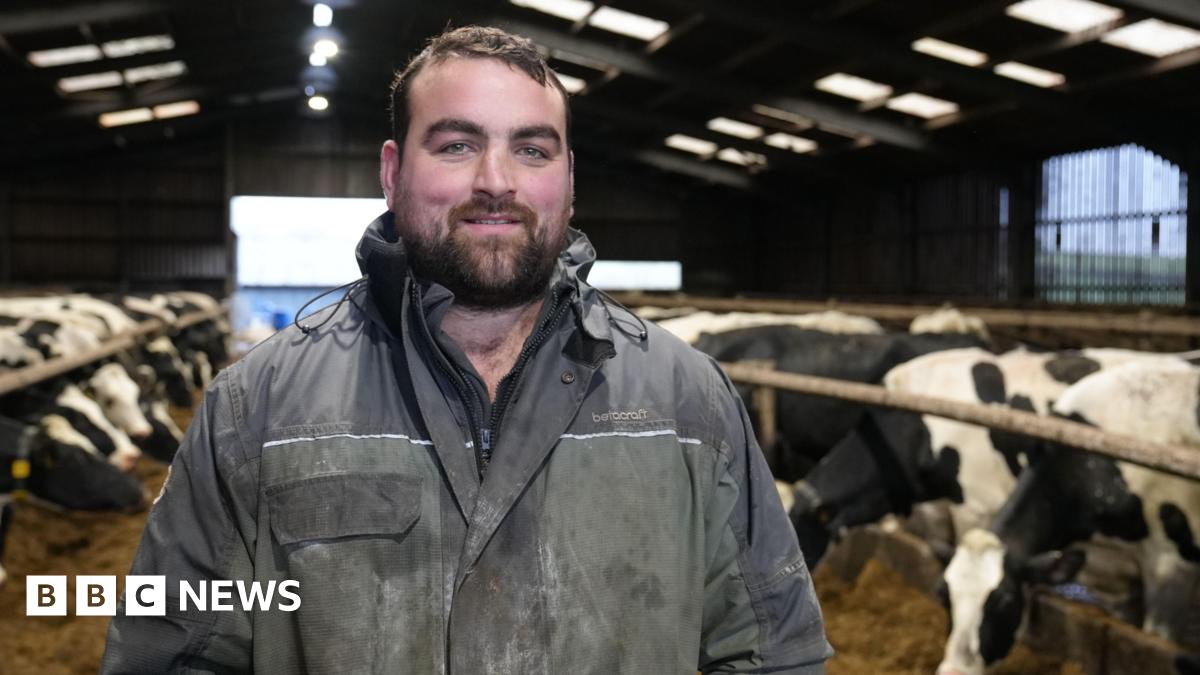 Gethin Hughes, dairy farmer stands in his cattle shed wearing blue overalls