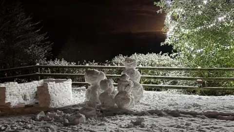 Emma Tsang Four snowmen and the start of an igloo in the garden of a house. With a green tree lit up in the background.