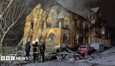 A smouldering residential building with a few firefighters standing out the front. There is lots of rubble around the building and covering a few cars that are parked out the front, and a bit of smoke coming from inside the building.