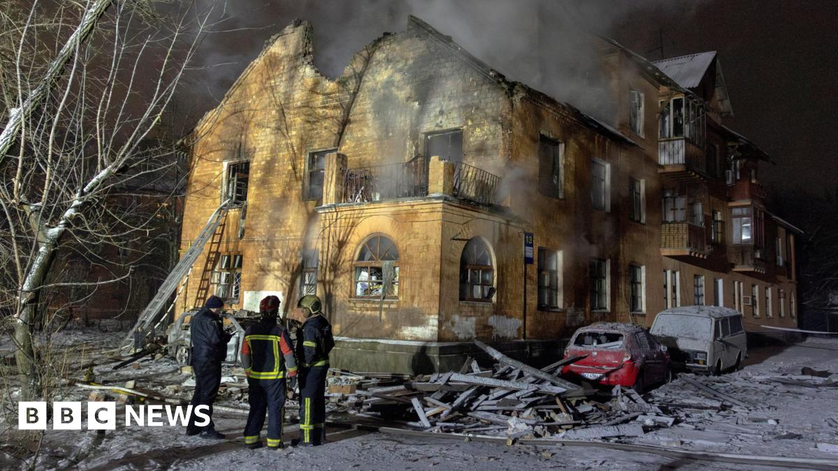 A smouldering residential building with a few firefighters standing out the front. There is lots of rubble around the building and covering a few cars that are parked out the front, and a bit of smoke coming from inside the building.