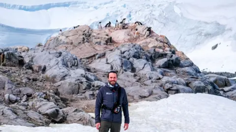 Tiago Figueiredo/UKAHT Pete Watson stands on snow looking at the camera. Behind him on a craggy rock are a dozen penguins with icebergs in the background.