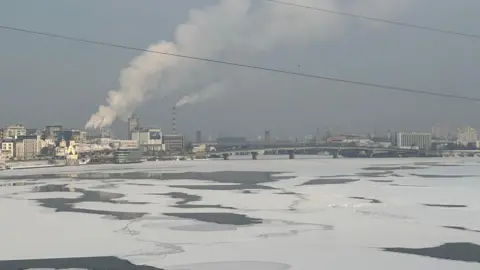 Scene of partly frozen Dnipro river in Kiev, with building and chimneys and a bridge in the background.
