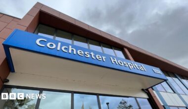 "Colchester Hospital" is written in white font on a blue banner above the hospital's entrance doors. The doors are glass, as is the whole facade.