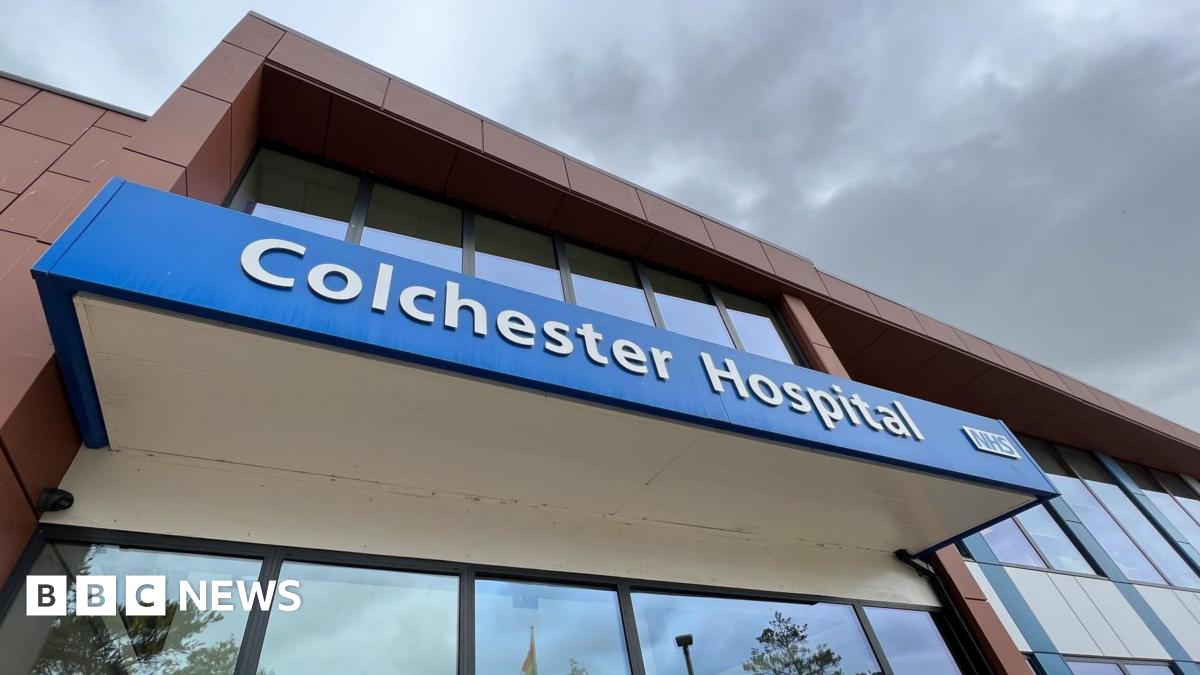 "Colchester Hospital" is written in white font on a blue banner above the hospital's entrance doors. The doors are glass, as is the whole facade.