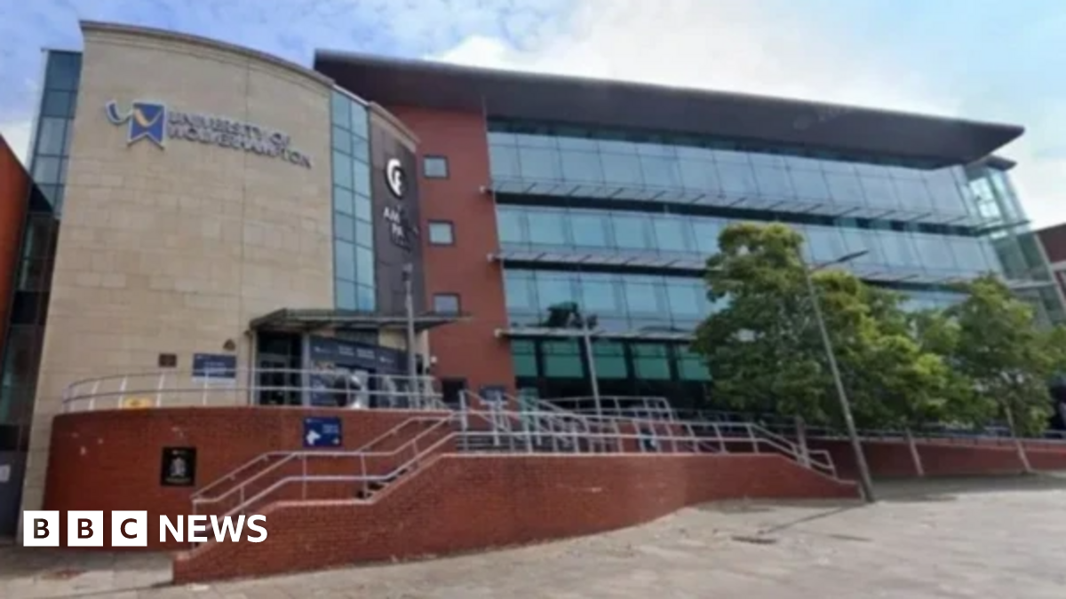 Millie Bobby Brown and David Harbour in character as Eleven and Jim Hopper. Eleven wears red shorts over grey leggings and a grey jumper. Jim Hopper wears a black leather jacket, a black beanie hat and dark trousers.They are speaking to director Frank Darabont on the set of Stranger Things. There is a vintage advertisement posted in the background with debrief on the ground.