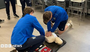 Millie Bobby Brown and David Harbour in character as Eleven and Jim Hopper. Eleven wears red shorts over grey leggings and a grey jumper. Jim Hopper wears a black leather jacket, a black beanie hat and dark trousers.They are speaking to director Frank Darabont on the set of Stranger Things. There is a vintage advertisement posted in the background with debrief on the ground.