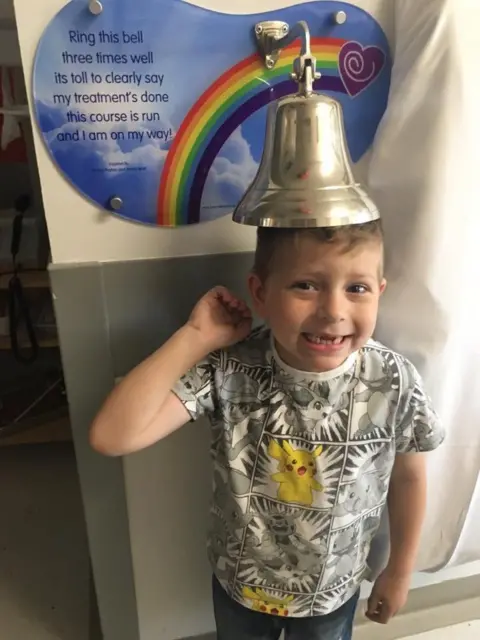 Tim Sadler Image of a young boy with gaps in his teeth smiling at the camera. He is standing under a brass ships bell. Behind the bell is a sign with blue skies, puffy clouds and a rainbow.