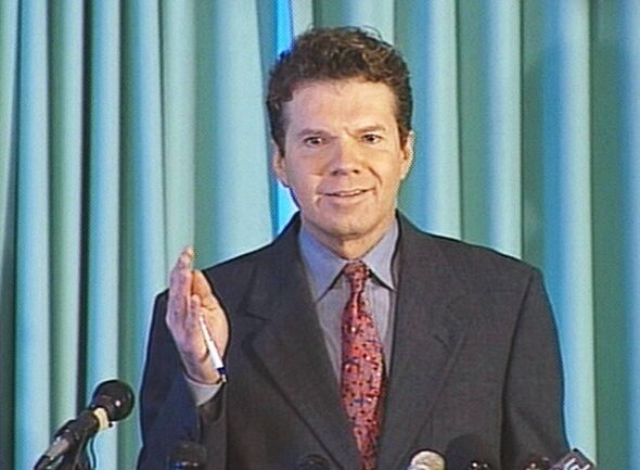 An individual dressed in a formal suit, consisting of a jacket and tie, is standing at a podium and gesturing with one hand whil