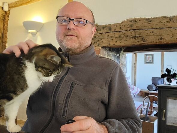 An individual wearing a gray turtleneck sweater, glasses, and holding a cat in their arms, standing in a room with wooden beams An individual wearing a gray turtleneck sweater, glasses, and holding a cat in their arms, standing in a room with wooden beams