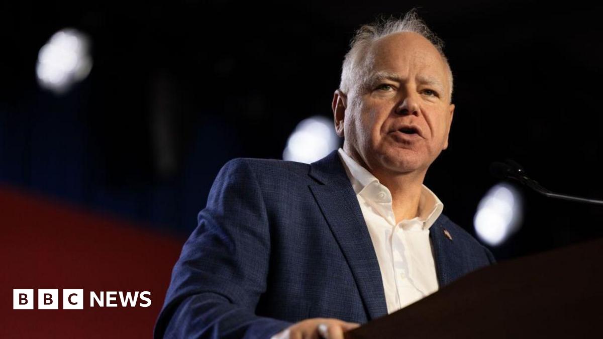 Tim Walz speaking at a podium wearing a dark blue suit jacket and a collared white shirt with no tie. He is mostly bald with some grey hair.