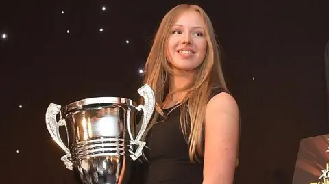 Maisy Creed Racing A girl with long hair wearing an evening dress holding a large trophy