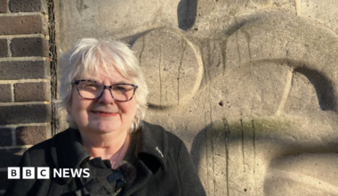 Angela Greenfield - a woman with short, grey hair, smiling. She is wearing dark glasses and a black coat. She is standing in front of a stone-coloured sculpture which is on the side of a car park