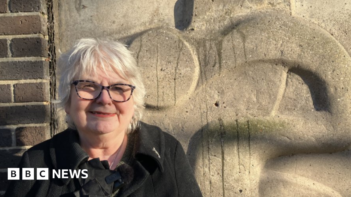 Angela Greenfield - a woman with short, grey hair, smiling. She is wearing dark glasses and a black coat. She is standing in front of a stone-coloured sculpture which is on the side of a car park