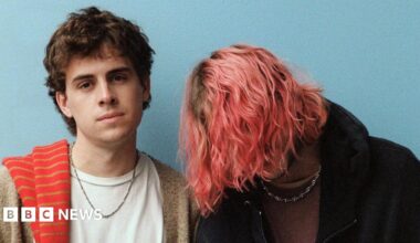 Press photo of Royel Otis standing against a plain blue background. Guitarist Royel Maddell covers his face with long, pink tousled hair