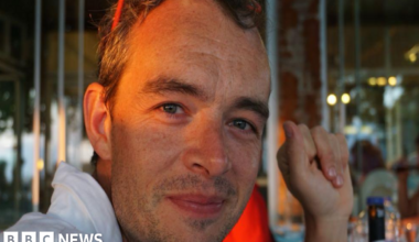 Marcus smiles into the camera, with evening light shining over his eyes. He's wearing a white shirt and has his arm resting on the table.