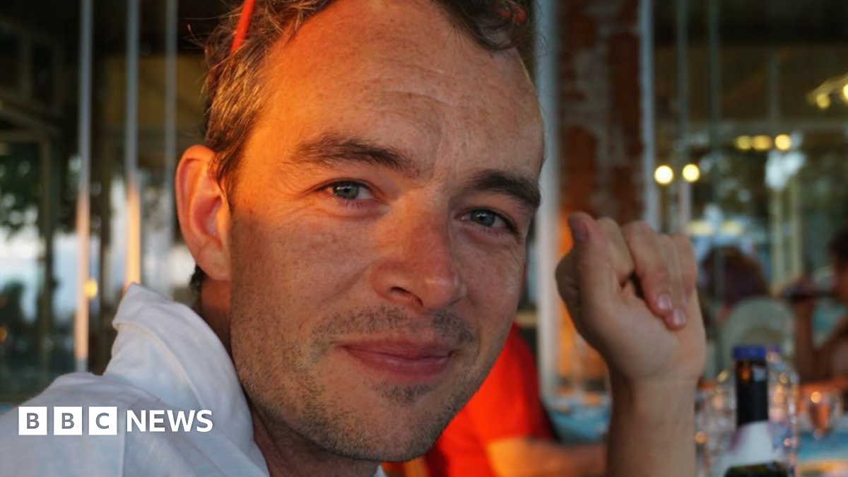 Marcus smiles into the camera, with evening light shining over his eyes. He's wearing a white shirt and has his arm resting on the table.