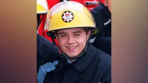 PA Media Actor John Alford playing the part of a firefighter: He is wearing  a yellow helmen, with a star-shaped badge, a blue coat and smiling.