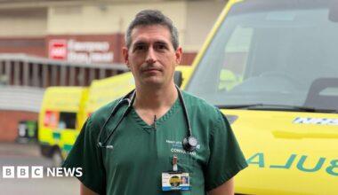 A man with dark grey hair stands in green hospital scrubs by an ambulance, in front of a hospital building.