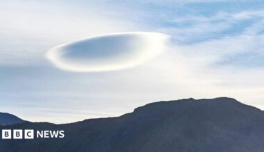 The picture shows a mountain on the beach, there is sand and a blue sky with 3 flat (UFO) clouds in the skyline.