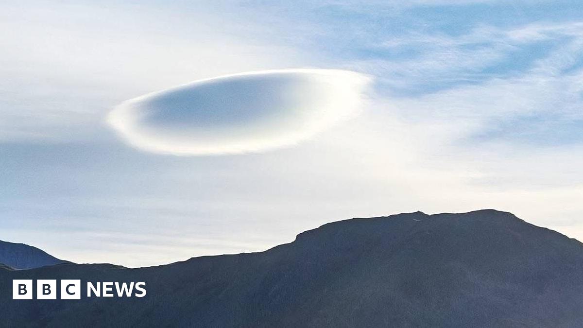 The picture shows a mountain on the beach, there is sand and a blue sky with 3 flat (UFO) clouds in the skyline.