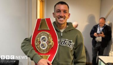 Millie Bobby Brown and David Harbour in character as Eleven and Jim Hopper. Eleven wears red shorts over grey leggings and a grey jumper. Jim Hopper wears a black leather jacket, a black beanie hat and dark trousers.They are speaking to director Frank Darabont on the set of Stranger Things. There is a vintage advertisement posted in the background with debrief on the ground.