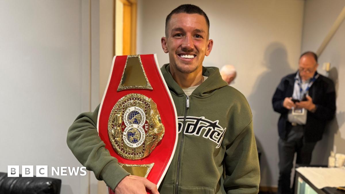 Millie Bobby Brown and David Harbour in character as Eleven and Jim Hopper. Eleven wears red shorts over grey leggings and a grey jumper. Jim Hopper wears a black leather jacket, a black beanie hat and dark trousers.They are speaking to director Frank Darabont on the set of Stranger Things. There is a vintage advertisement posted in the background with debrief on the ground.