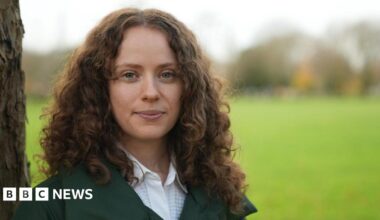 Holly stares to camera. She is in focus with dark, long curly hair and is leaning against a tree-trunk while the background is out of focus but looks like a green field.