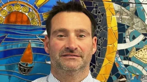 Tom Lewis Headshot of Tom Lewis. He is white with short brown hair and a stubbly beard growth. The collar of a white and grey-striped shirt is visible. Behind him is a mosaic mural of a sea with a boat on it, with the year 2010 on it, and abstract patterns and a fish tail to one side.