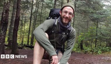 Alex Pretti smiles and kneels on one knee in a wooded area. He wears a green hat, green long-sleeved top, brown shorts, and a black hiking backpack. He has brown facial hair. It is daytime.