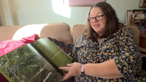 BBC Jane Bertelli, a woman in her mid sixties, sits on the sofa in her home, looking through one of the brochures sent to her by Ethical Forestry Limited.
