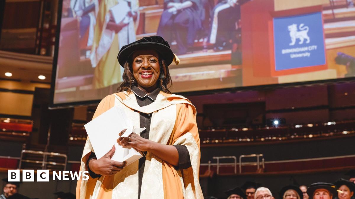 Marverine Cole stands on a stage in a graduation gown and hat, holding her doctorate. She smiles at the camera. Her robe is yellow and white.