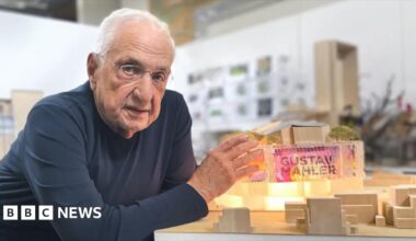 A person wearing a dark long-sleeve shirt is leaning on a table, presenting a detailed architectural model, featuring illuminated sections, miniature trees, and geometric structures in a modern studio setting.