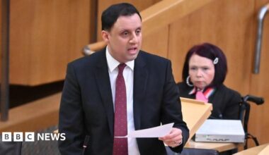 Prime Minister Mark Carney, wearing a black suit jacket, and a white button-up shirt leans his chin on his hands. He has short grey and black hair and is looking away from the camera.