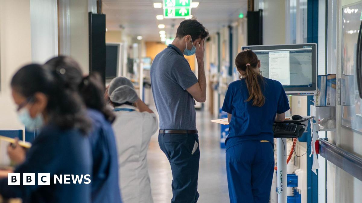Generic view of two medical staff members from behind looking at a laptop. Some other people in scrubs are seen blurred walking around them.