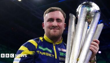 Luke Littler celebrates with the Sid Waddell trophy after victory against Gian van Veen after the final of the World Darts Championship at the Alexandra Palace. He is wearing his signature purple and yellow darts shirt and smiling.