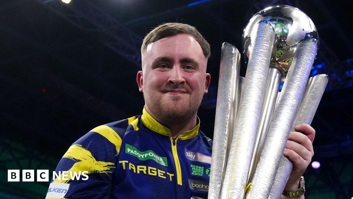 Luke Littler celebrates with the Sid Waddell trophy after victory against Gian van Veen after the final of the World Darts Championship at the Alexandra Palace. He is wearing his signature purple and yellow darts shirt and smiling.