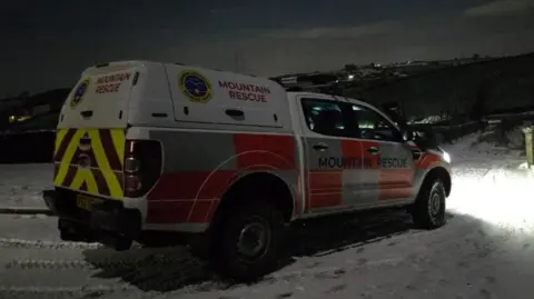Mourne Mountain Rescue Team A Mourne Mountain Rescue Team vehicle parked in the snow in the dark. The 4X4 is white with red and yellow high viz stripes and silver reflective panels. The team's name and logo is on the sides and rear of the 4X4