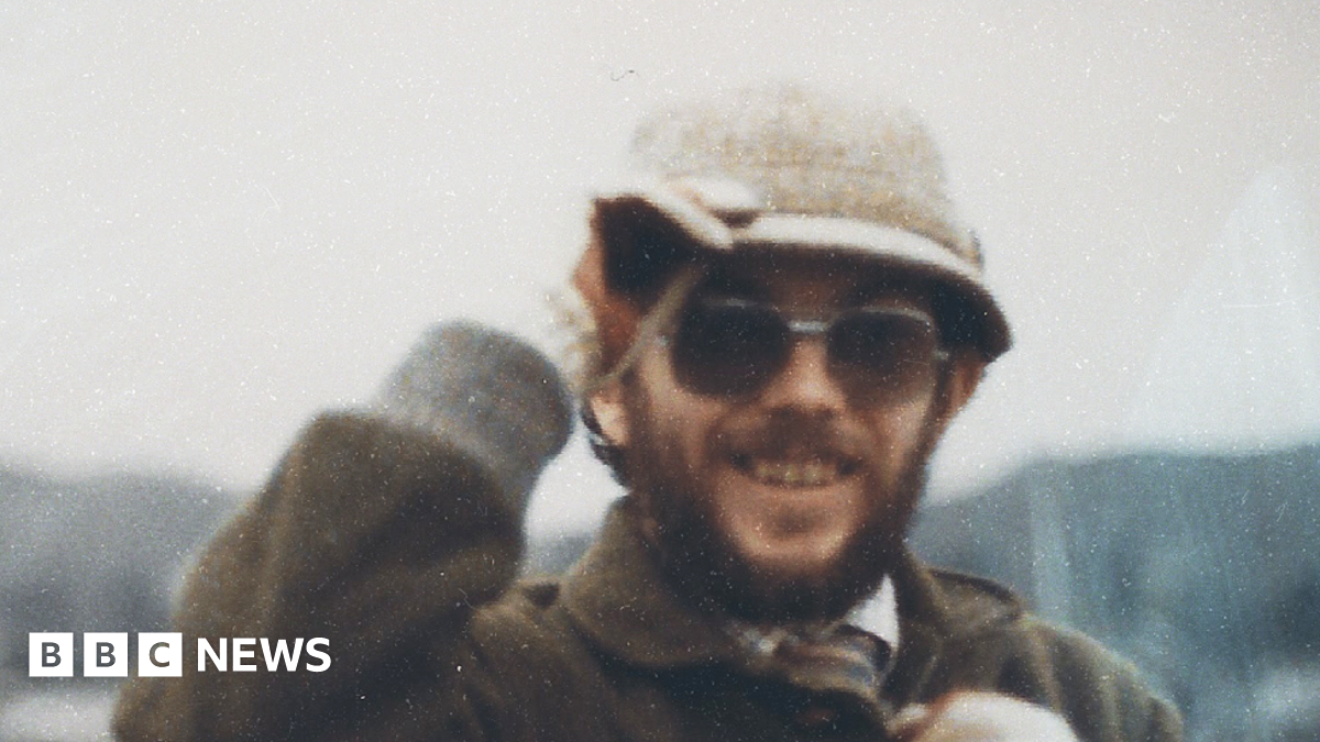 Kenner Elias Jones is a white man in his 30s or 40s in this picture, with a brown beard and moustache. He wears dark lensed prescription glasses and a cream coloured hat which he is holding, a checked shirt and tie covered by a brown coat with a collar. He is smiling at the camera. The image is grainy.