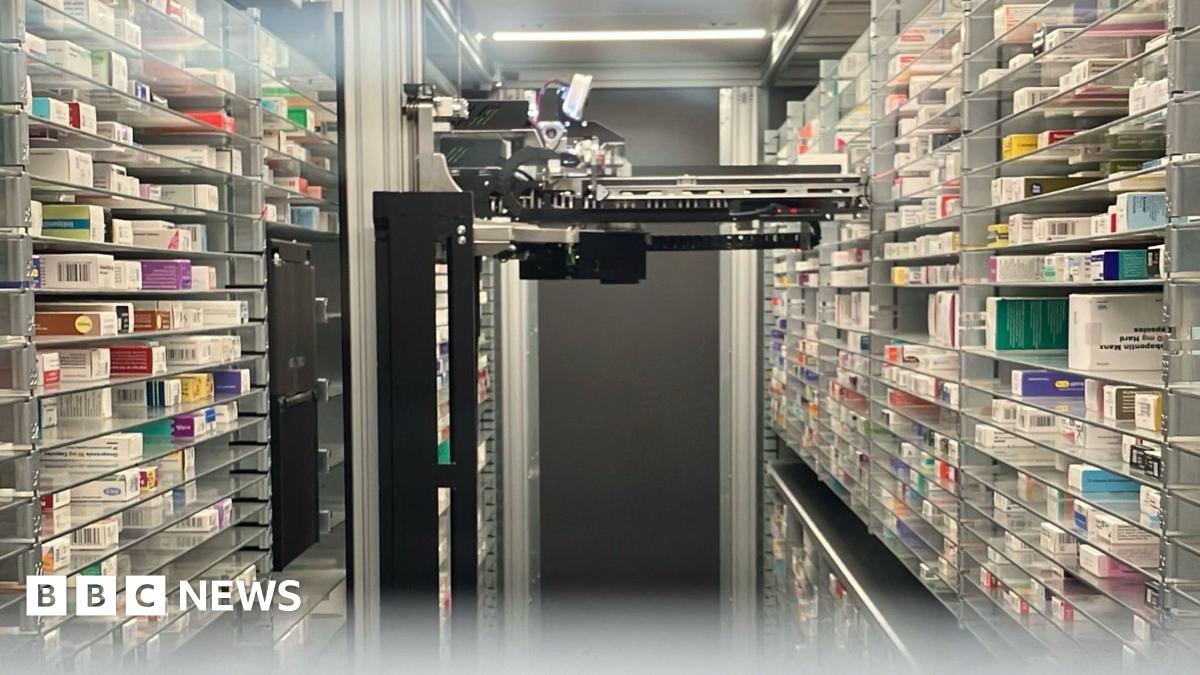 A black robotic device is positioned in the middle of a room. On either side are shelves stacked with various types of medication. There is a dark grey door at the far end of the room behind the robot.