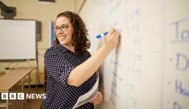 A happy curly brown haired female teacher writing on whiteboard in classroom. She has a navy polka dot shirt on