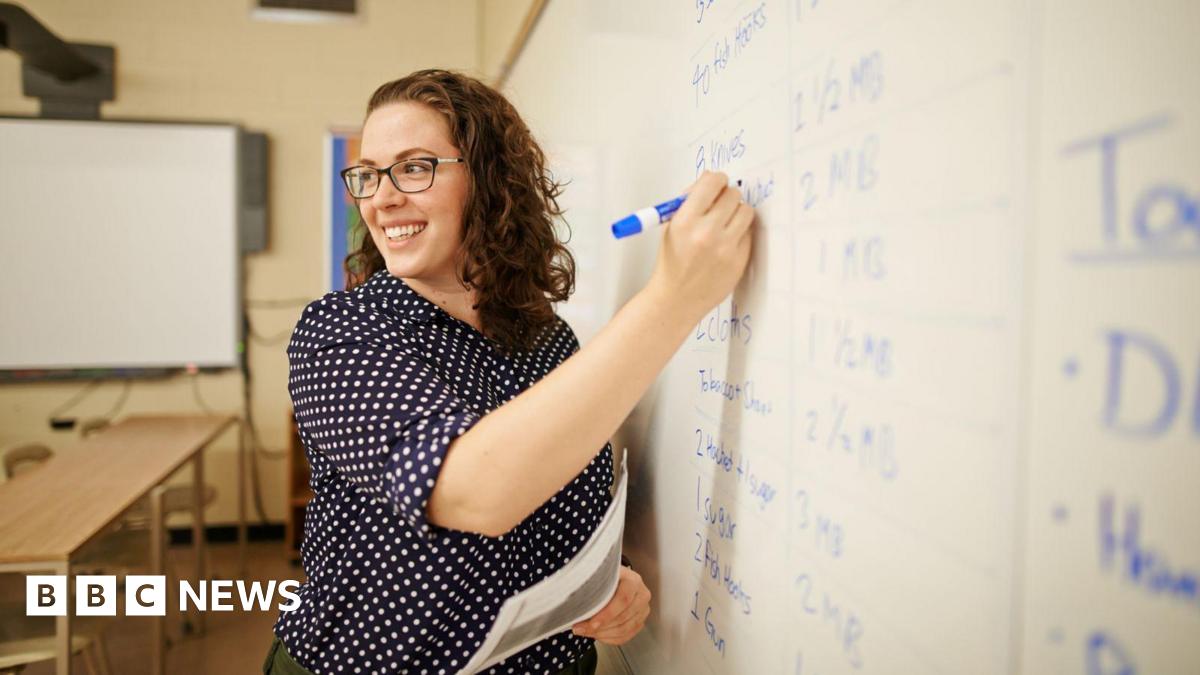 A happy curly brown haired female teacher writing on whiteboard in classroom. She has a navy polka dot shirt on