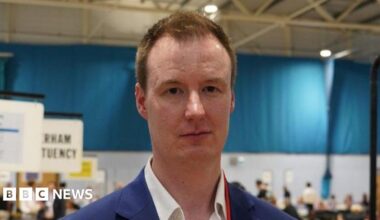A line of police in riot gear stand across a road as fireworks are launched at them.