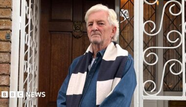 Anthony Burton, a man with white hair and a dark denim shirt, stands outside his home looking disgruntled.