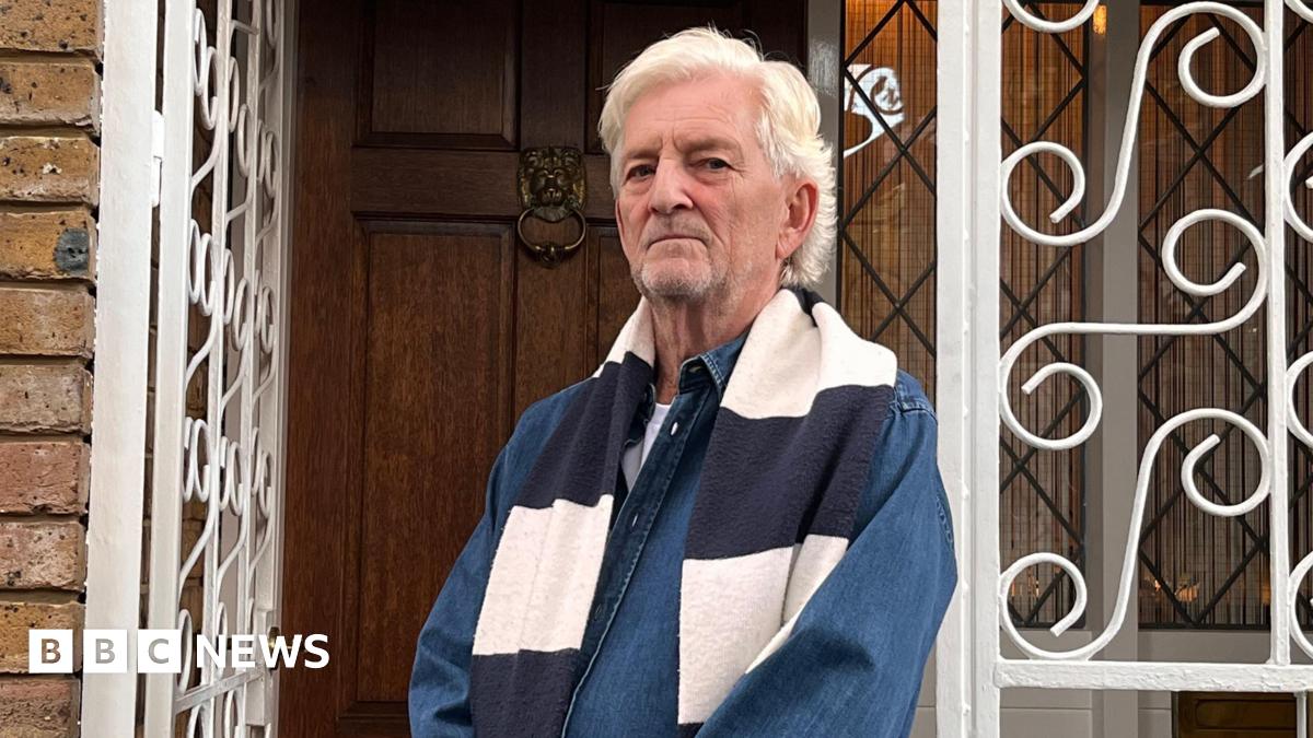 Anthony Burton, a man with white hair and a dark denim shirt, stands outside his home looking disgruntled.