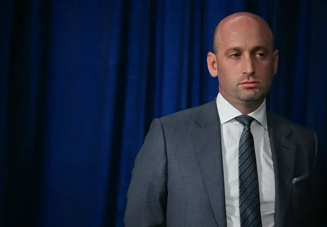 Stephen Miller stands in a suit during a press conference with a blue backdrop behind him